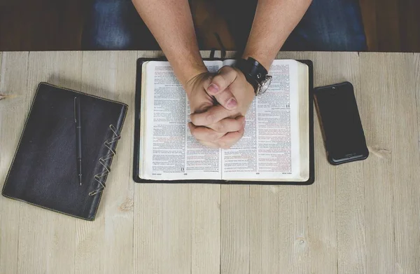 Overhead tiro de um macho com a mão na bíblia orando perto de um caderno e uma caneta — Fotografia de Stock