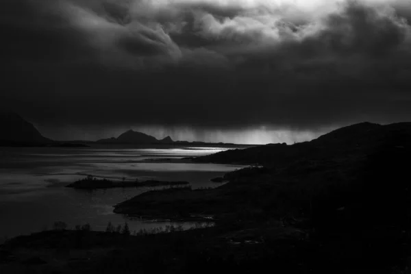 Tepelerin siluetlerinin gri ölçekli görüntüsü ve gökyüzünün altında kara bulutlu bir göl. — Stok fotoğraf