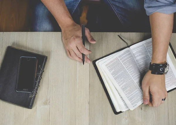 Overhead shot of a male reading the bible and holding a pen near his notebook and smartphone