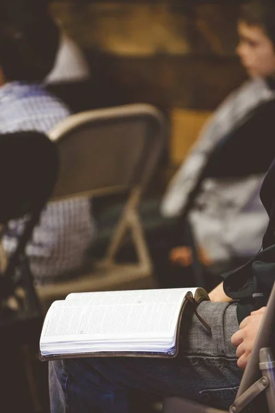 Vertikale Aufnahme eines Mannes, der mit einer offenen Bibel auf dem Oberschenkel vor verschwommenem Hintergrund sitzt — Stockfoto