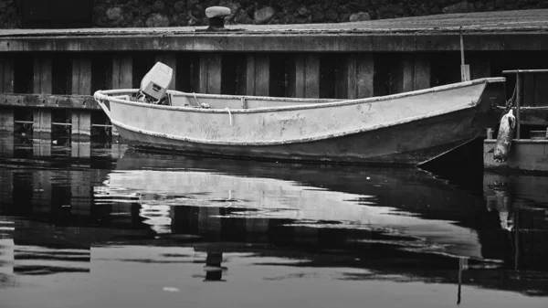 A grayscale shot of a rusty boat reflected in the lake near a pier