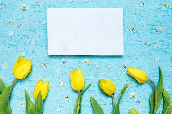 Tiro de ângulo alto de um papel em branco e tulipas amarelas em uma superfície azul com pequenas flores de margarida — Fotografia de Stock
