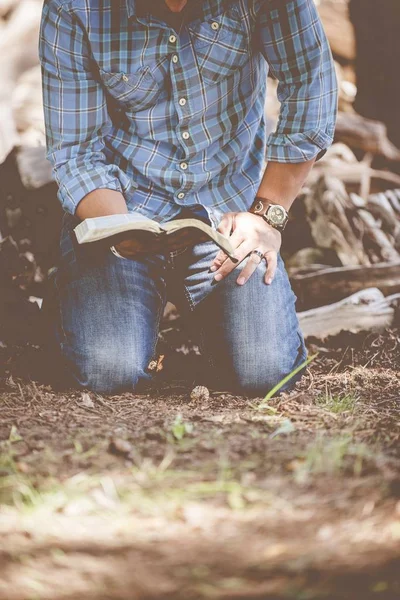 A vertical shot of a male in a casual clothing kneeled on the soil reading the holy bible