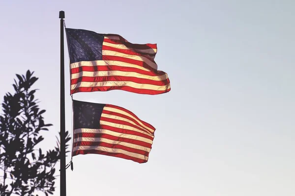 Two American flags waving on a single metal pole on a bright sunny day