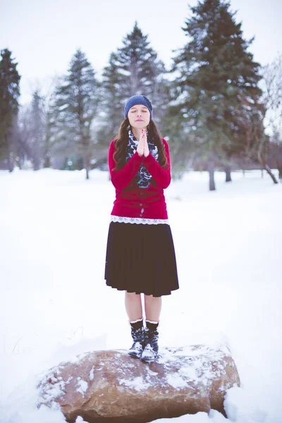 赤いジャケットを着た黒髪の女性と 雪の領域で目を閉じて祈る石の上に青い帽子 — ストック写真