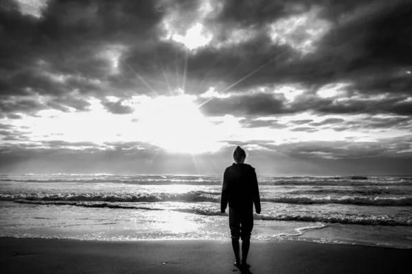 Scatto in scala di grigi di una femmina in piedi sulla spiaggia con la luce del sole nel cielo nuvoloso — Foto Stock