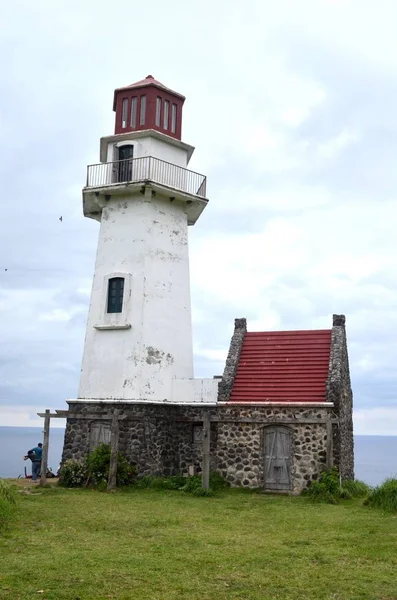 Белый маяк с красной крышей в окружении зелени с морем и рыбаками на заднем плане — стоковое фото