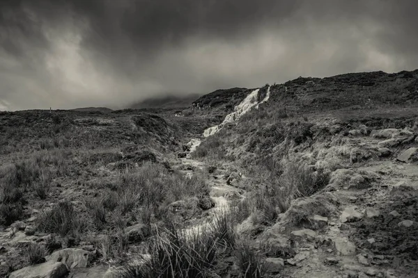 Escala de grises de un valle con un río rodeado de piedras y hierba bajo un cielo gris oscuro — Foto de Stock