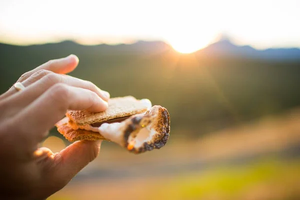 Person holding s'more made with marshmallow chocolate and cookies with the sunset in the background — Stock Photo, Image
