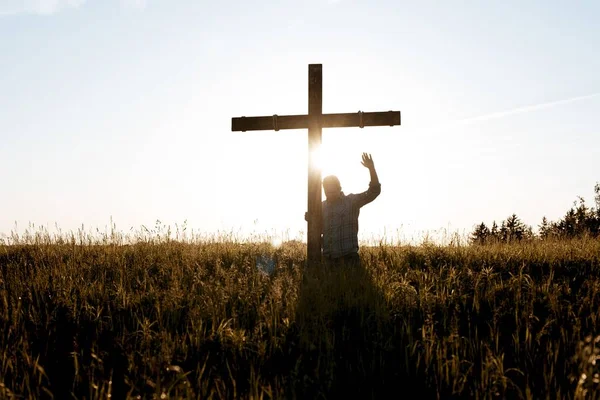 Man staat bij een handgemaakt houten kruis met zijn hand omhoog naar de hemel biddend — Stockfoto