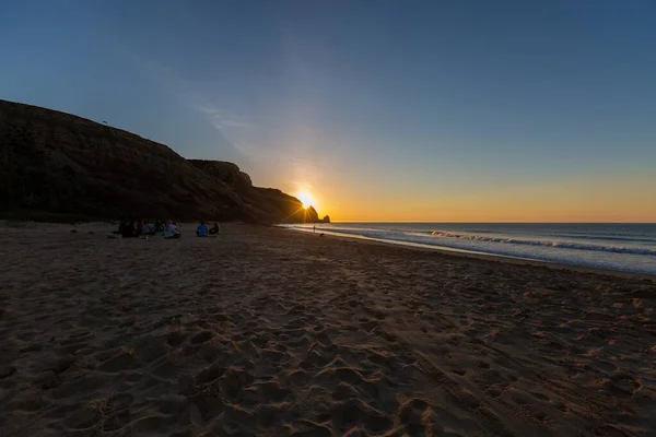 Cenário de uma praia com pessoas sentadas nela rodeadas por rochas e mar durante o pôr-do-sol em Portugal — Fotografia de Stock