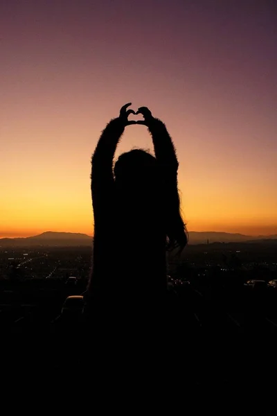 Tiro vertical de una silueta de una hembra haciendo una forma de corazón con sus manos durante la puesta del sol — Foto de Stock