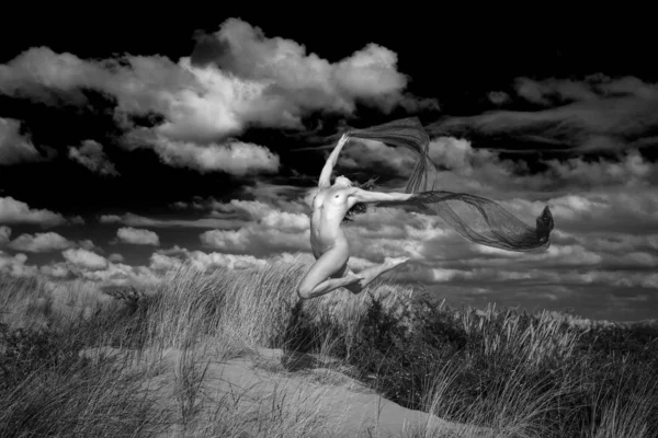 Grayscale shot of a naked woman in the state of a jump in the air with a black texture in her hands — Stockfoto