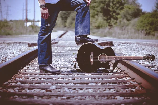 Shallow focus shot of a male wearing boots and standing on train tracks with one foot on guitar — 스톡 사진