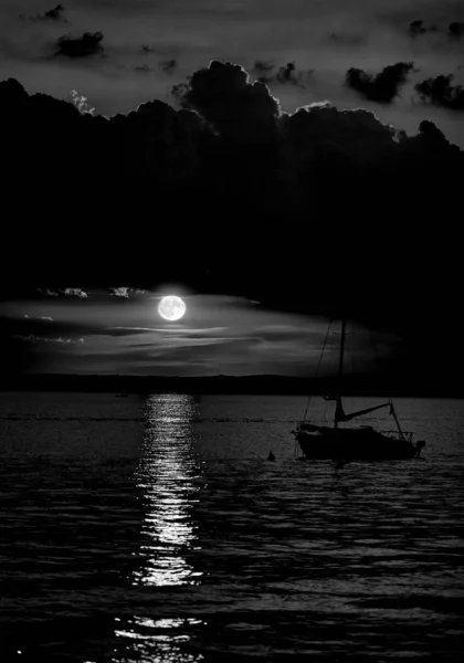 Lanzamiento a escala de grises de un barco en el mar bajo el oscuro cielo nublado con la luna al fondo — Foto de Stock