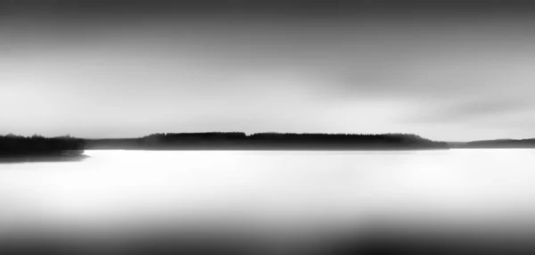 Greyscale panoramic view of a sea under a cloudy sky with a forest behind the fog — ストック写真