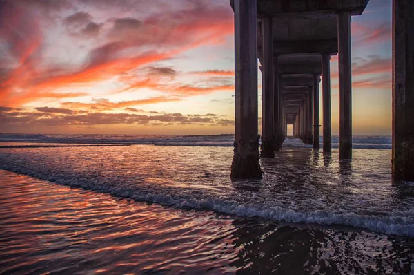 Okyanus dalgalarının alçak açılı görüntüsü sıcak renkli gökyüzünün altındaki köprüye sıçrıyor. — Stok fotoğraf