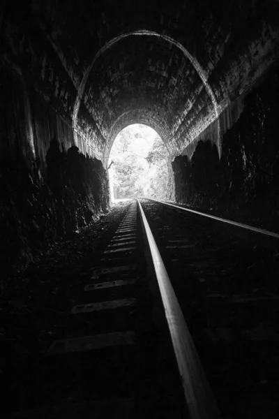 Vue verticale en échelle de gris d'un chemin de fer vide dans un tunnel avec une lumière vive qui brille à la fin — Photo
