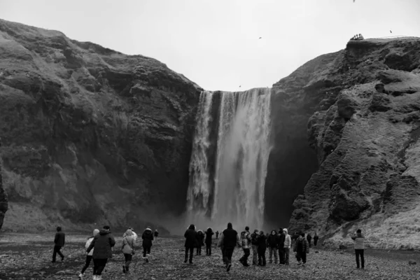 Black and white shot of a group of tourists taking photographs of the tall waterfall in Iceland — Stock Photo, Image