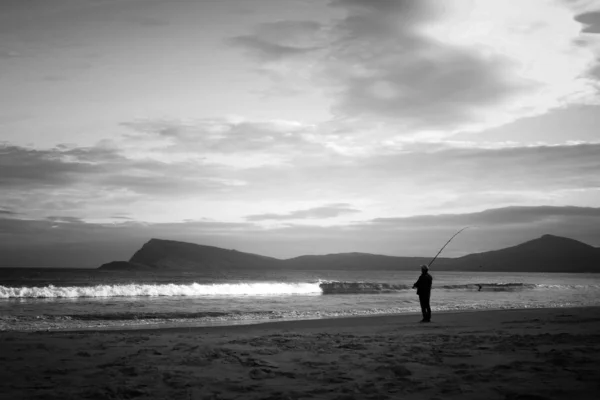 Серый Снимок Человека Пляже Удочкой Гористыми Холмами Вдалеке Облачным Небом — стоковое фото