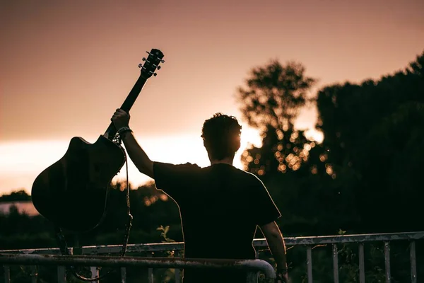 Man Holding Guitar Metallic Fences Greenery Blurry Background Sunset — 스톡 사진