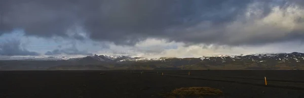 Panoramic shot of an empty field with snowy mountains in the distance under a cloudy sky — 스톡 사진