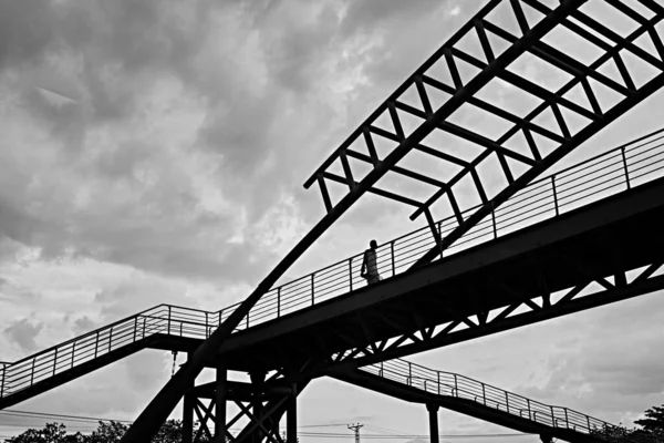 Scala di grigi colpo basso angolo di un maschio che cammina attraverso un ponte sotto il cielo nuvoloso — Foto Stock