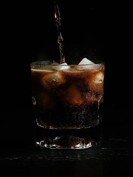 Verticaal schot cokes dat wordt gegoten in een glas vol ijs op een zwarte achtergrond — Stockfoto