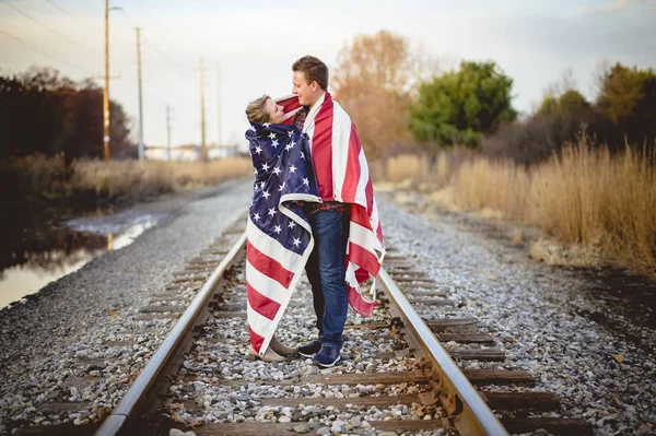Young couple with the American flag around their shoulders standing on the railway — Stockfoto