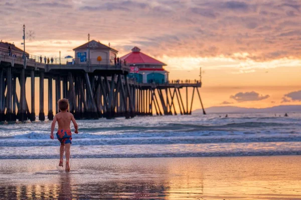 Boy running through the beach surrounded by buildings and the sea during the sunset — Stok fotoğraf