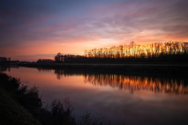 Silhouettes of trees under the cloudy sunset sky reflected in the lake below — 스톡 사진