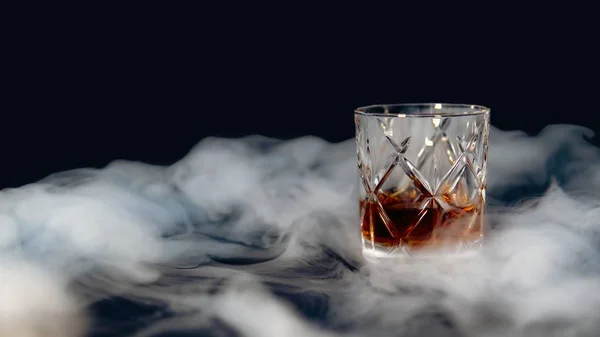 Vidro de uísque em uma mesa coberta de fumaça contra um fundo preto — Fotografia de Stock