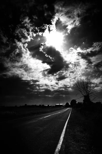 Scatto verticale in scala di grigi del sole che si nasconde dietro le nuvole di tempesta su una strada vuota — Foto Stock