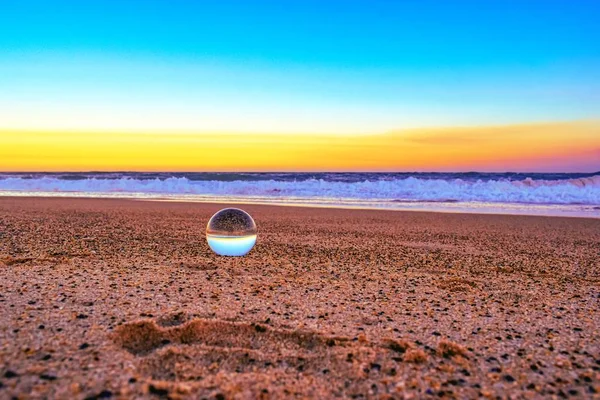 Primer plano de una bola transparente en la arena rodeada por el mar durante la puesta del sol en la noche — Foto de Stock