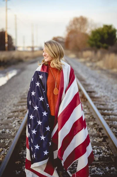 Vertical shot of a female with the American flag on her shoulders standing on the railway — Stock Photo, Image