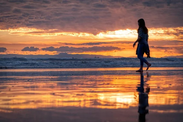 Chica paseando por la playa rodeada por el mar bajo un cielo nublado durante una puesta de sol dorada. —  Fotos de Stock