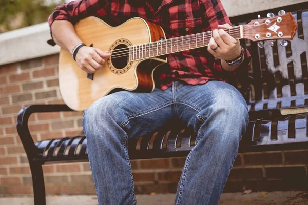 Kırmızı ve siyah flanel giyen bir erkek bankta oturmuş gitar çalıyor. — Stok fotoğraf