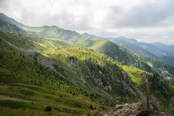 Krajina kopců pokrytá zelení s skalnatými horami pod zataženou oblohou na pozadí — Stock fotografie