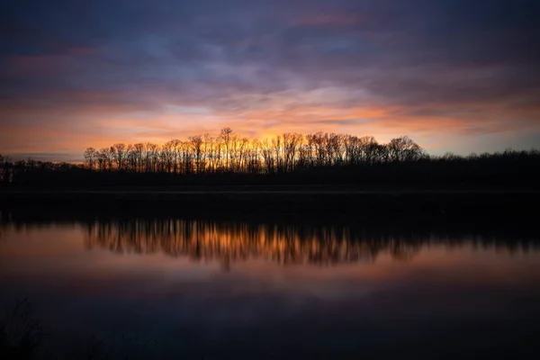 Siluetas de árboles bajo el cielo nublado del atardecer reflejado en el lago debajo —  Fotos de Stock