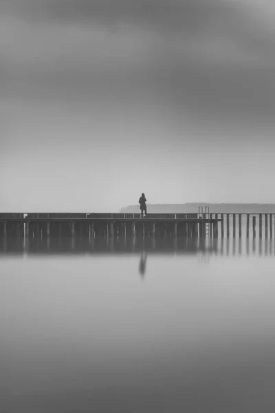 Vertical greyscale shot of a lonely person standing on a wooden dock near the sea — Stock Photo, Image