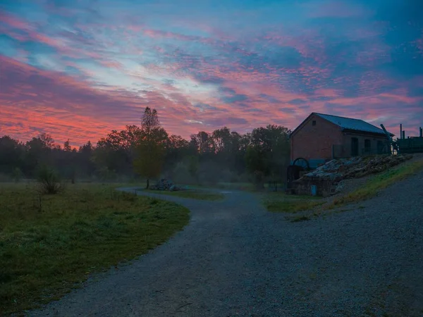 A view of two-way road surrounded by greenery with a cottage and a colorful sunset in the background