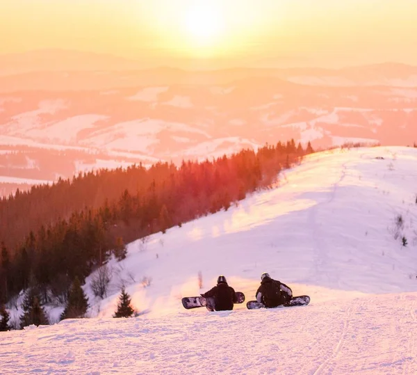 Високий кут зйомки двох лижників, які сидять на яскравому снігу, спостерігаючи за заходом сонця з лісом на відстані — стокове фото
