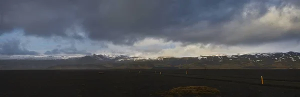 Panoramic shot of an empty field with snowy mountains in the distance under a cloudy sky — 스톡 사진