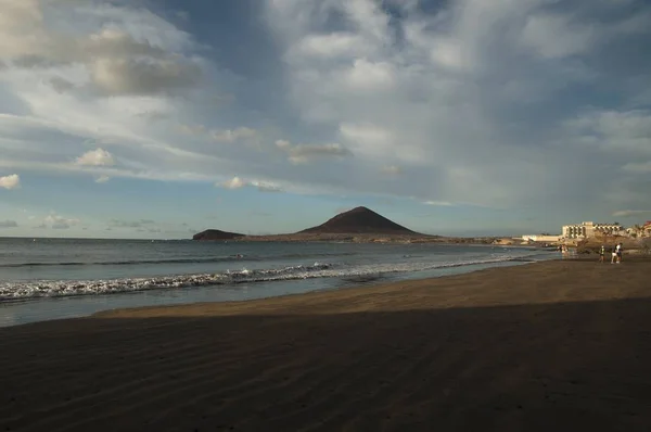 Beach surrounded by people and the sea with hills under a cloudy sky and sunlight on the background — Stok fotoğraf