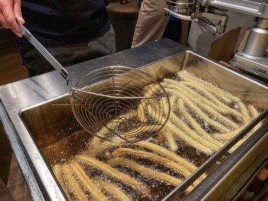 A mans hand cooking churros in cooking in oil, close up of a hand holding a strainer taking cooked churros out of boiling oil clipart