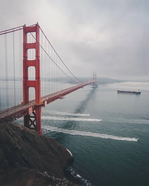 Tiro vertical da ponte portão dourado com um navio navegando à distância sob um céu nublado — Fotografia de Stock