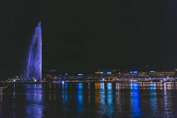 Широкоугольный снимок фонтана в окружении зданий и воды в ночное время — стоковое фото