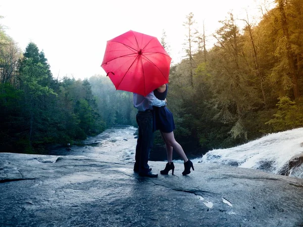 Junto con un paraguas rojo besando en un parque rodeado de vegetación y cascada. — Foto de Stock