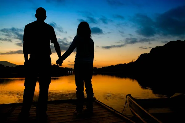 Siluet dari pasangan berpegangan tangan di jembatan kayu yang dikelilingi oleh danau selama matahari terbenam — Stok Foto