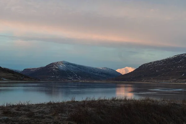雪に覆われた丘に囲まれた湖は、アイスランドの日没時の水を反映しています。 — ストック写真